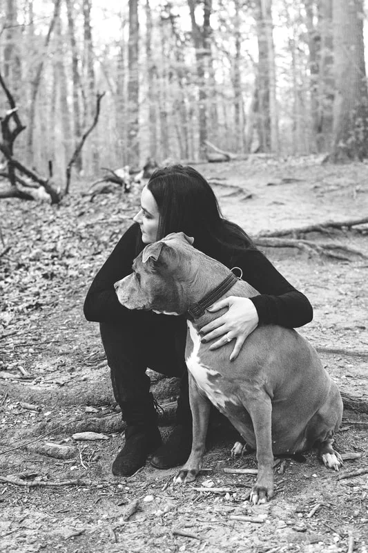 A black and white photo of a pit bull and her owner sitting together looking to the side in a forest