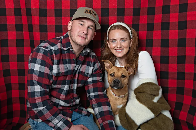Color photo of a light brown tripod three legged dog against a buffalo plaid background with its family. The photo was taken by Laura Pea Photography, a pet photographer on Long Island.