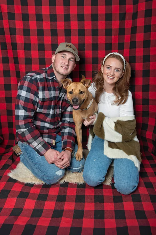 Color photo of a light brown tripod three legged dog with her family posing against a buffalo plaid background. The photo was taken by Laura Pea Photography, a pet photographer on Long Island.