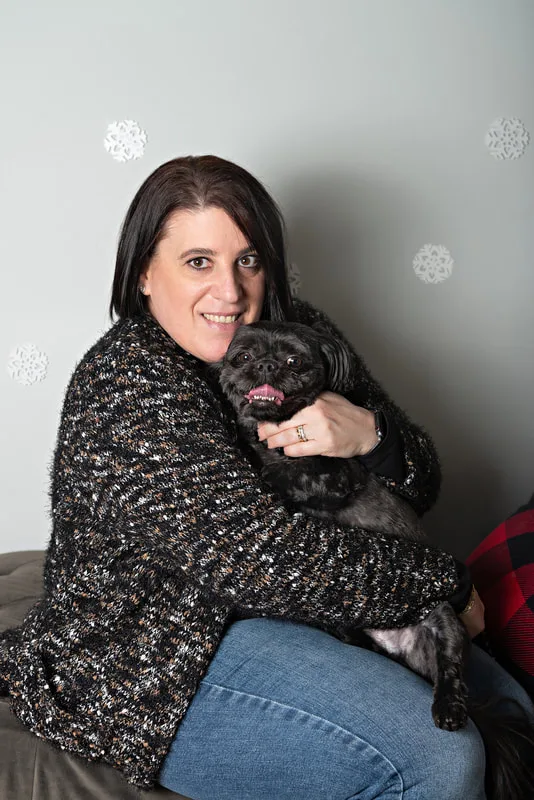 Color photo of a black shih tzu breed dog sitting on a grey couch in front of a snowflake background on his owner's lap, a white woman with brown hair. The photo was taken by Laura Pea Photography, a pet photographer based on Long Island, NY. 