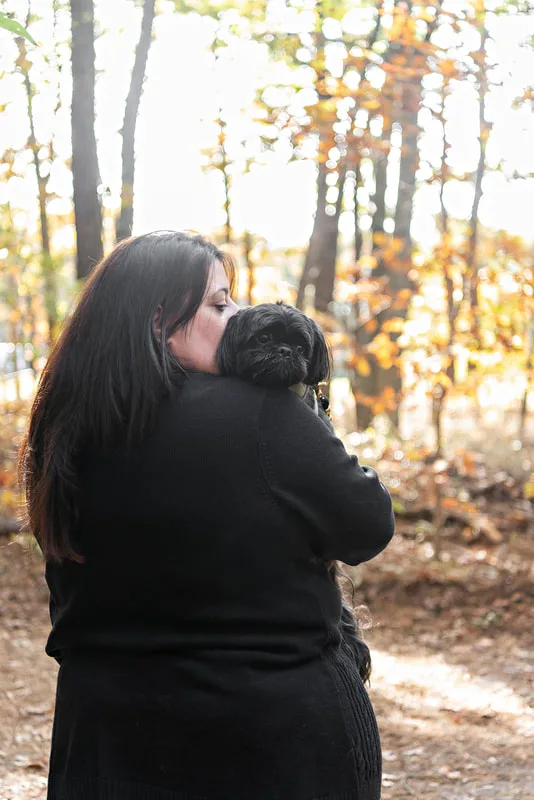 photo of a black shih tzu dog in the woods with his mom who is kissing him on the head
