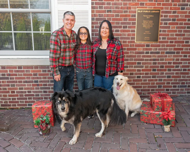 Color photo of a mom, dad and tween boy with their two husky mid dogs in front of red wrapped gifts and Christmas props. The photo was taken by Laura Paesano of Laura Pea Photography, a Long Island pet photographer. 