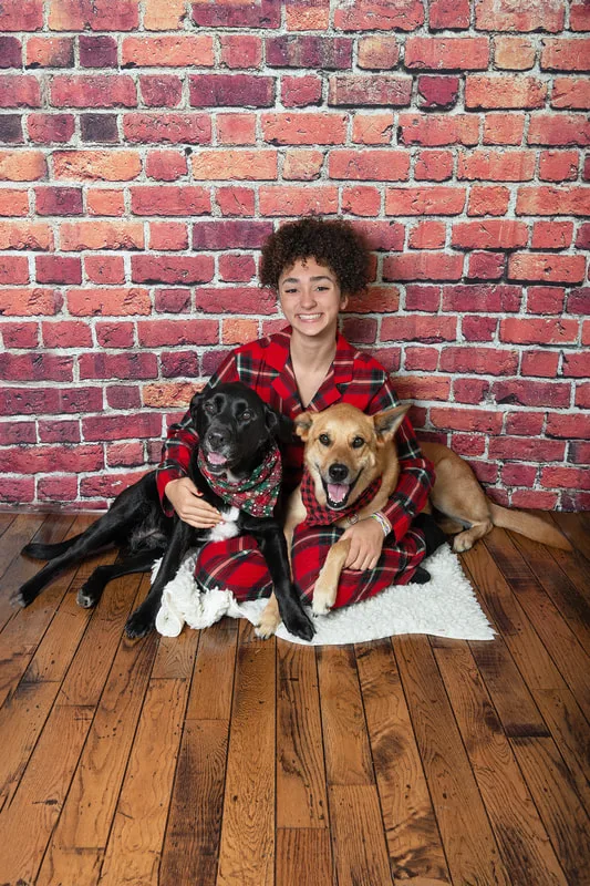 Color photo of a teenage girl wearing Christmas pajamas with her two dogs, a black labrador retriever mix and a fawn shepherd mix, taken in a photo studio against a red brick background. The photo was taken by Laura Pea Photography, a Long Island pet photographer. 