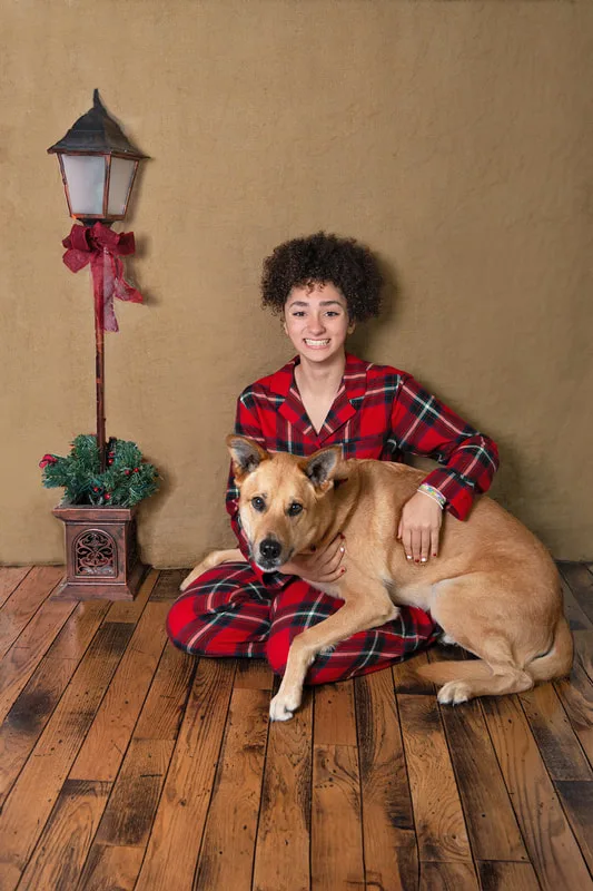 Color photo of a teenage girl wearing Christmas pajamas sitting on a wood floor with her fawn shepherd mix dog, taken in a photo studio against a brown burlap background. The photo was taken by Laura Pea Photography, a Long Island pet photographer. 