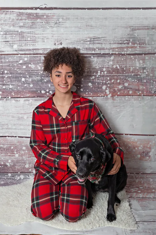 Color photo of a teenage girl wearing Christmas pajamas sitting on a white furry rug with her black labrador retriever mix dog, taken in a photo studio against a snowy wood background. The photo was taken by Laura Pea Photography, a Long Island pet photographer. 