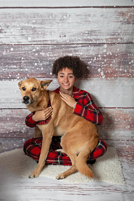 Color photo of a teenage girl wearing Christmas pajamas sitting on a white furry rug with her fawn shepherd mix dog, taken in a photo studio against a snowy wood background. The photo was taken by Laura Pea Photography, a Long Island pet photographer. 