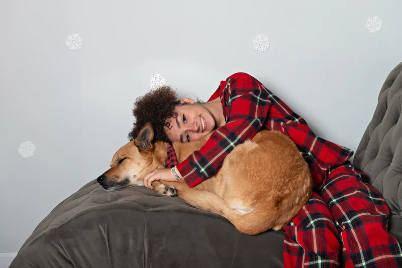 Color photo of a teenage girl wearing Christmas pajamas sitting on a grey couch with her \fawn shepherd mix dog, taken in a photo studio against a snowflake background. The photo was taken by Laura Pea Photography, a Long Island pet photographer. 