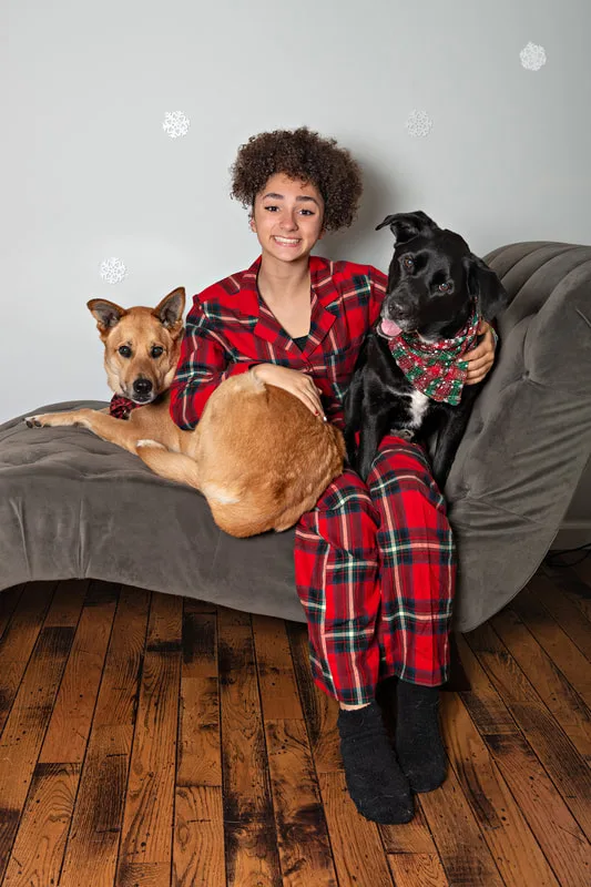 Color photo of a teenage girl wearing Christmas pajamas sitting on a grey couch with her two dogs, a black labrador retriever mix and a fawn shepherd mix, taken in a photo studio against a snowflake background. The photo was taken by Laura Pea Photography, a Long Island pet photographer. 