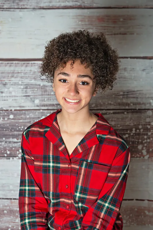 Color photo of a teenage girl wearing Christmas pajamas. taken in a photo studio against a snowy wood background. The photo was taken by Laura Pea Photography, a Long Island pet photographer. 
