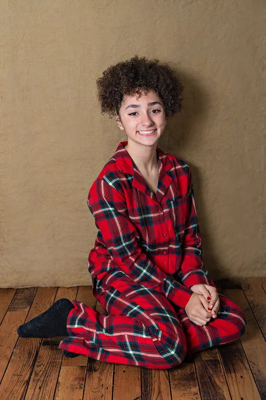 Color photo of a teenage girl wearing Christmas pajamas sitting on a wood floor taken in a photo studio against a brown burlap background. The photo was taken by Laura Pea Photography, a Long Island pet photographer. 