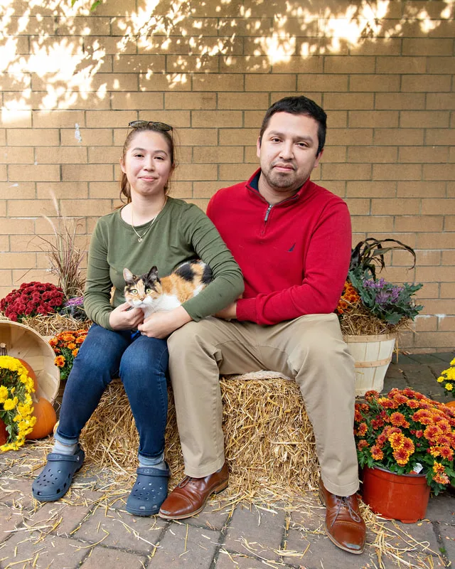 color photo of a fall display with hay bales, mums, pumpkins and two people with their cat