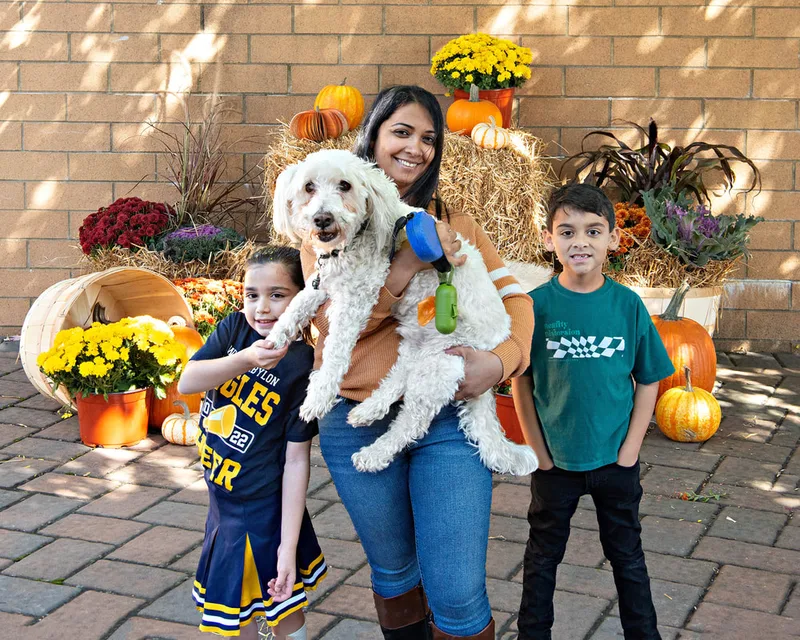 color photo of a fall display with hay bales, mums, pumpkins and a mother with her two children and small dog