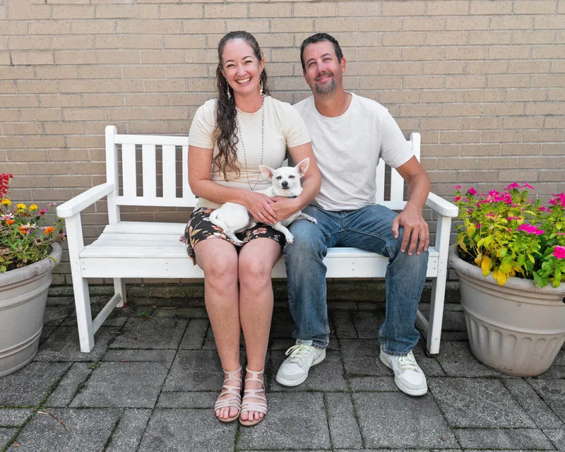 color photo of a Chihuahua dog with her owners on a white bench next to flowers. The photo was taken at West Islip Public Library by Long Island Pet Photographer Laura Pea Photography. 