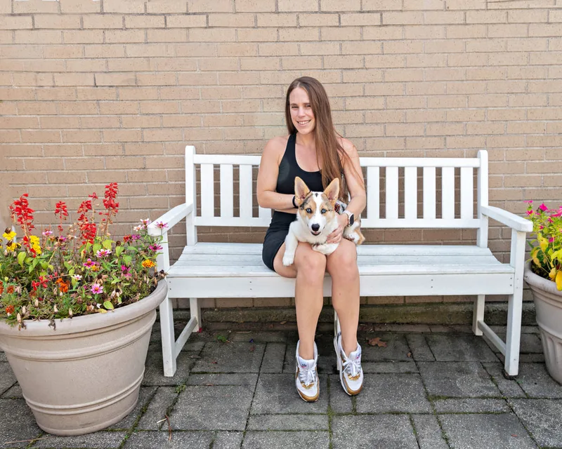 color photo of a corgi dog and owner on a white bench next to flowers. The photo was taken at West Islip Public Library by Long Island Pet Photographer Laura Pea Photography. 