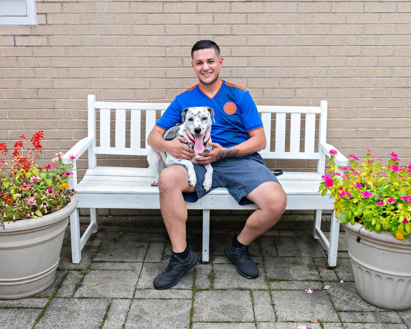 color photo of a dalmatian mix dog and owner on a white bench next to flowers. The photo was taken at West Islip Public Library by Long Island Pet Photographer Laura Pea Photography. 