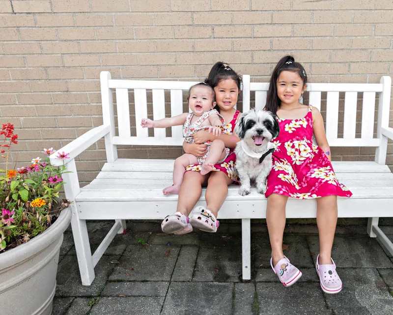 color photo of a black and white dog with three little girls on a white bench next to flowers. The photo was taken at West Islip Public Library by Long Island Pet Photographer Laura Pea Photography. 