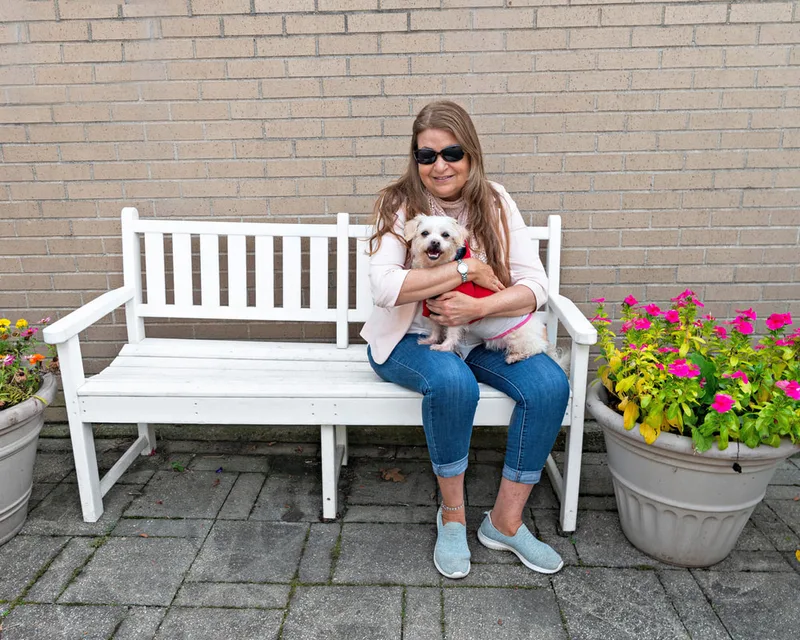 color photo of a terrier dog and owner on a white bench next to flowers. The photo was taken at West Islip Public Library by Long Island Pet Photographer Laura Pea Photography. 
