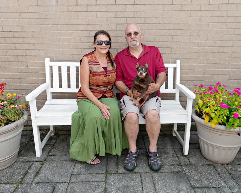 color photo of a miniature pinscher dog and owners on a white bench next to flowers. The photo was taken at West Islip Public Library by Long Island Pet Photographer Laura Pea Photography. 