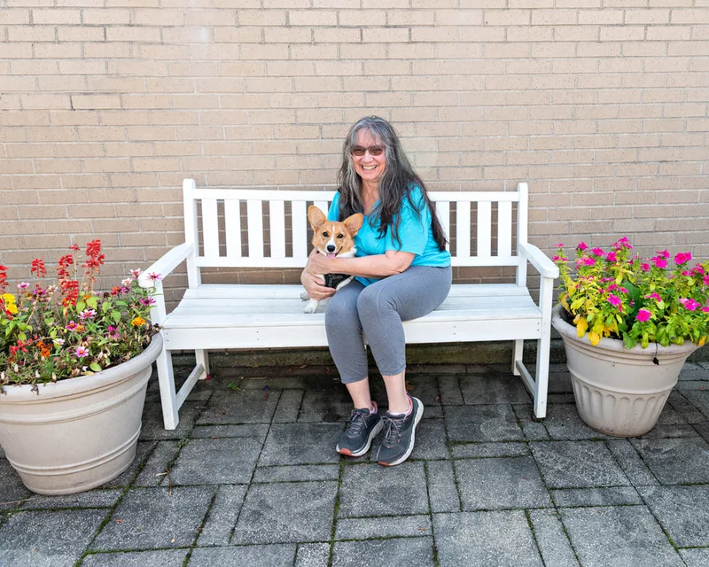 color photo of a corgi dog and owner on a white bench next to flowers. The photo was taken at West Islip Public Library by Long Island Pet Photographer Laura Pea Photography. 