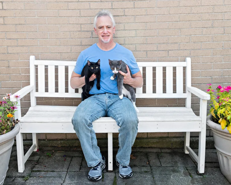 color photo of a man with two cats on a white bench next to flowers. The photo was taken at West Islip Public Library by Long Island Pet Photographer Laura Pea Photography. 