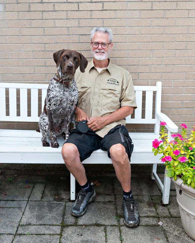 color photo of a pointer dog and owner on a white bench next to flowers. The photo was taken at West Islip Public Library by Long Island Pet Photographer Laura Pea Photography. 