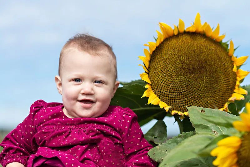 one year old baby girl smiling next to a sunflower on long island. Photo taken by Laura Pea Photography