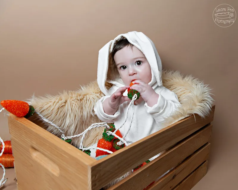 picture of baby boy's first easter photo session in an easter bunny costume sitting in a wooden crate chewing on carrot shaped lights