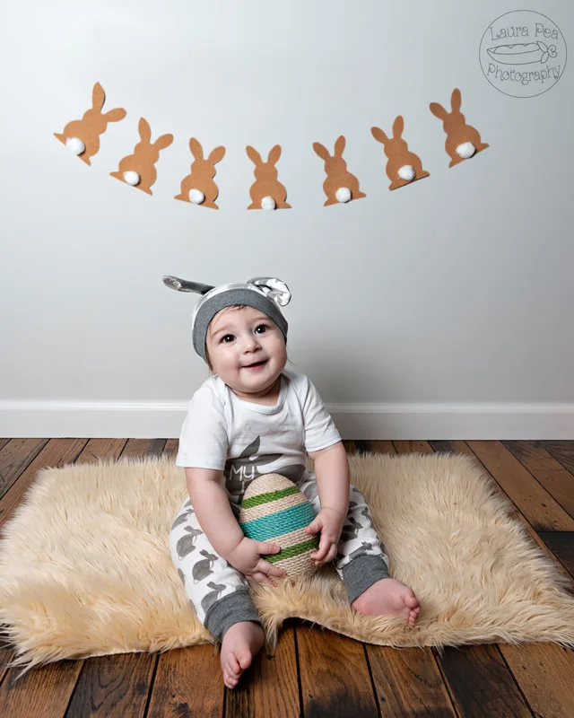 picture of baby boy's first easter photo session in an easter bunny costume smiling at the camera holding a twine easter egg