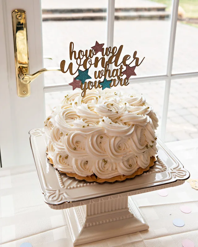 picture of a vanilla frosted cake with a golden cake topper that reads how i wonder what you are