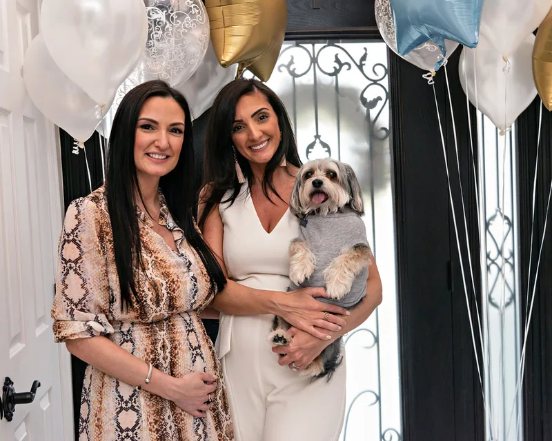 picture of two pregnant sisters holding their family dog in a hallway at their gender reveal party