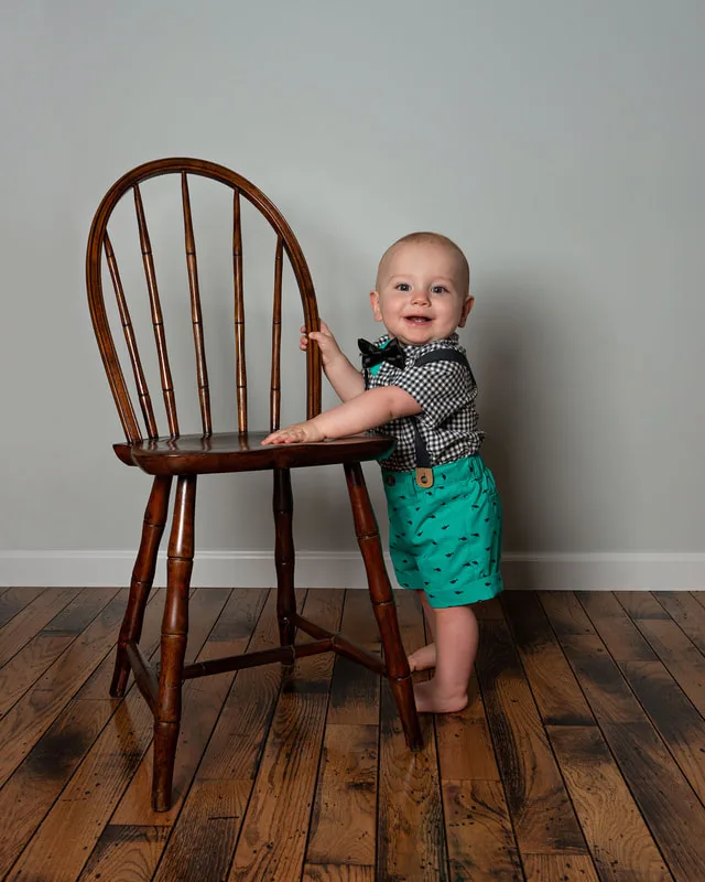 One year old boy first birthday photo in plaid outfit standing holding onto a chair