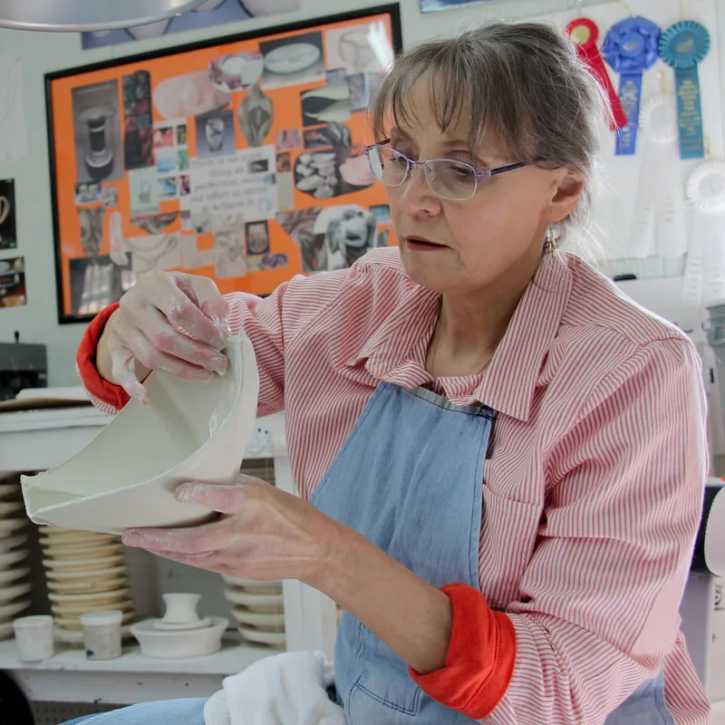 Antoinette Badenhorst shows how she alters a wheelthrown porcelain bowl. Behind her are several awards that show her achievements in pottery.  