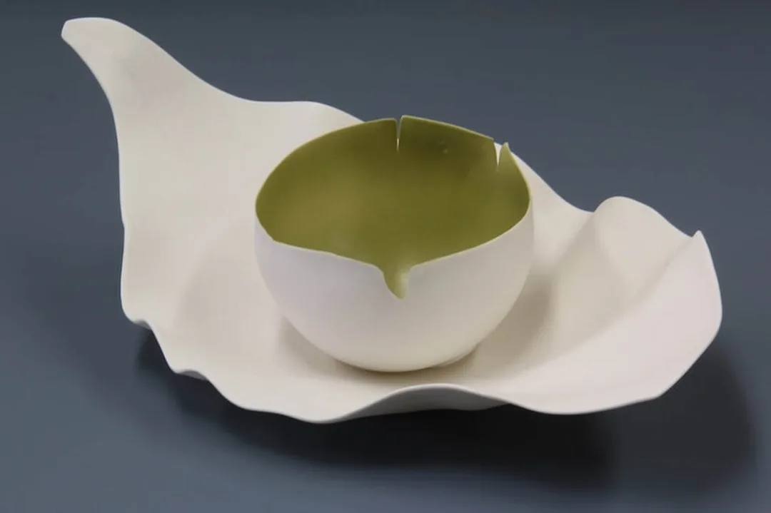 A hand-built porcelain sculpture by Antoinette Badenhorst featuring two parts. A small, round, white bowl with a green interior and a jagged rim sits within a larger, flat, white porcelain sheet that resembles a leaf or a large petal. The bowl has a small V-shaped opening on one side. 