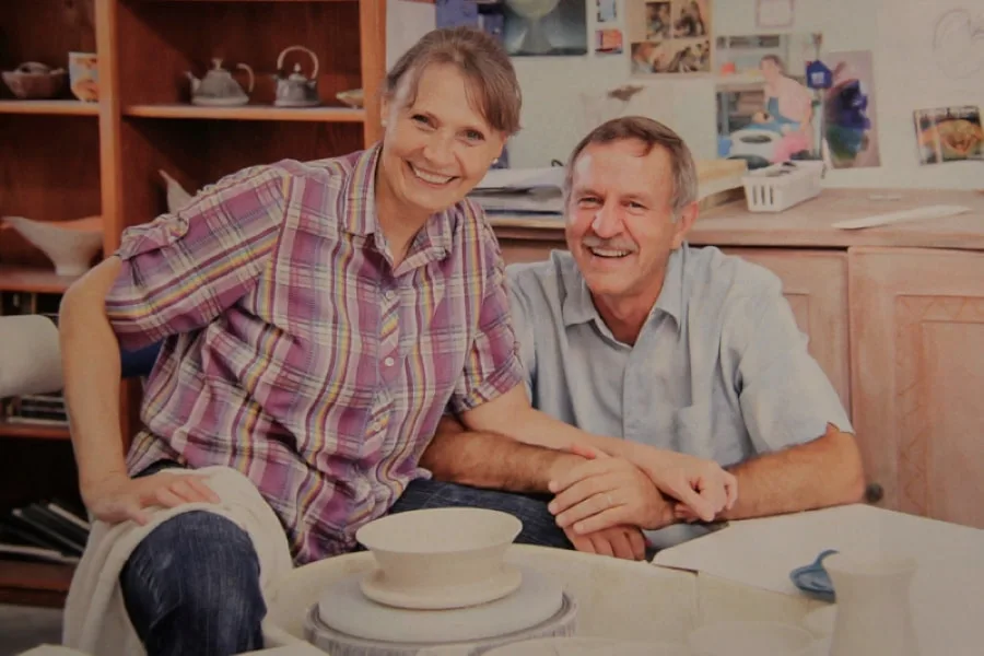 Koos and Antoinette Badenhorst during an interview with Mud & Magnolia magazine in the pottery studio of Antoinette. They are sitting at the pottery wheel and several of Antoinette's translucent porcelain bowls are visible on the shelf behind her.  