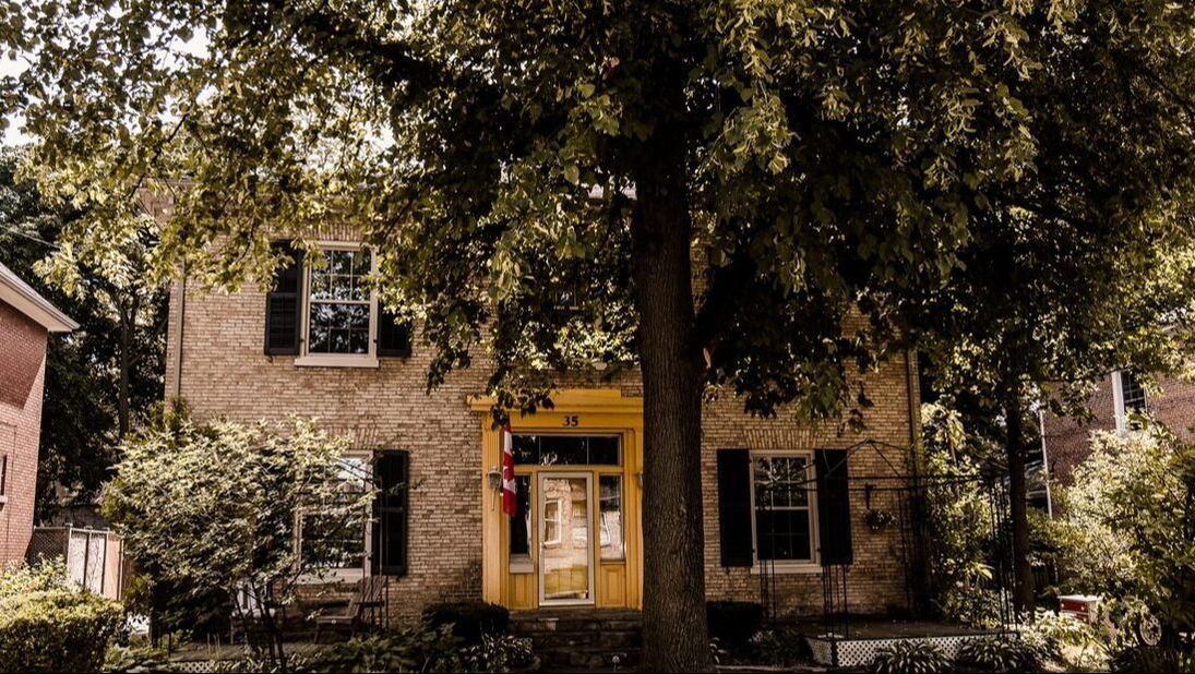 Here is an image of a historical home in St. Thomas. The house has a bright yellow front entrance. There is a tree standing just off centre of the shot. The house is covered in beiege and white brick and has black shutters. 