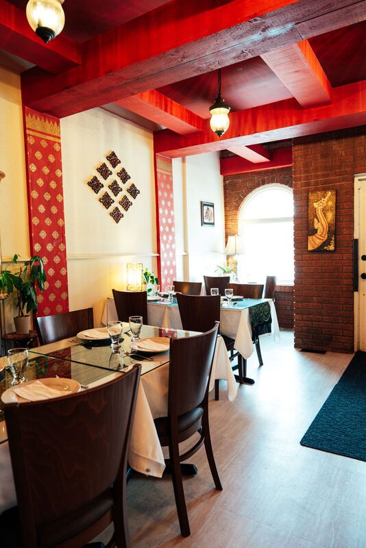 Here is Lotus Thai restaurant. Straight ahead is the historic beige bricked building. The front door is red with windows on either side. The windows are also framed in red. The second floor of the house has three windows, with the one in the middle having a black balcony bar in front. Below the house is a small front deck with a ramp to the right. In front of the deck is a table of four sitting on the patio. At the very front of the picture is a sign that says 'Lotus Thai Parking in Rear' with an arrow pointing left.