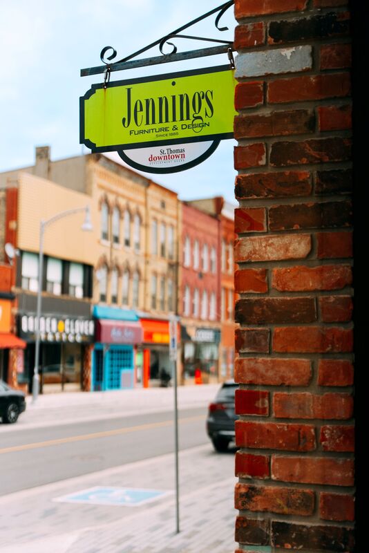 This is the store front of Mugford's. There is an orange and red awning above the store windows. The sign has an orange background and in the bottom left corner of it, it says 