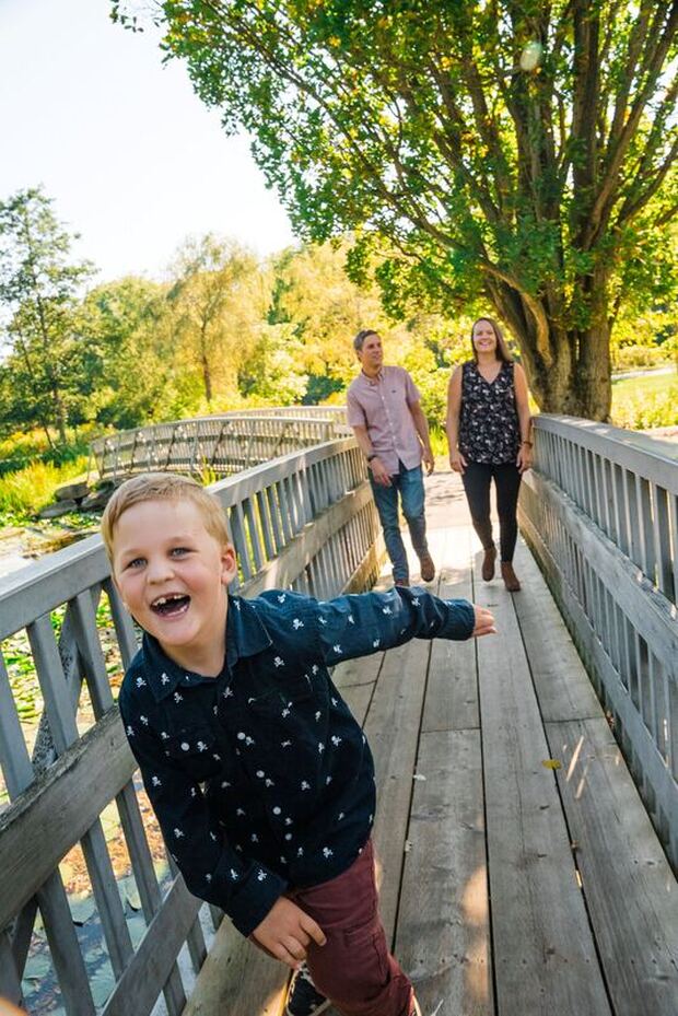 Here is a man and two children crossing one of the bridges at Waterworks Park. The girl is wearing a white and red checkered dress. The man is wearing a black shirt with sunglasses. In front is a boy wearing a blue and white patterned shirt. In the back are many trees.