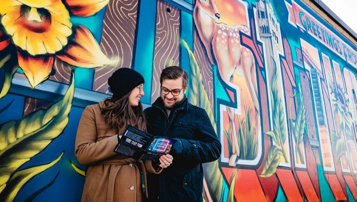 Here is an image of the boxcars by the Elgin County Railway Museum. To the left is the Anything Is Possible mural. The background is of the Anything Is Possible boxcar mural. It shows a woman with blue hair, a green polka dot shirt, red scarf and pink skin. She is blowing bubbles. On the right is the TRIL/RAPIDÉ mura; which is an abstract piece with white, orange, red and dark grey.