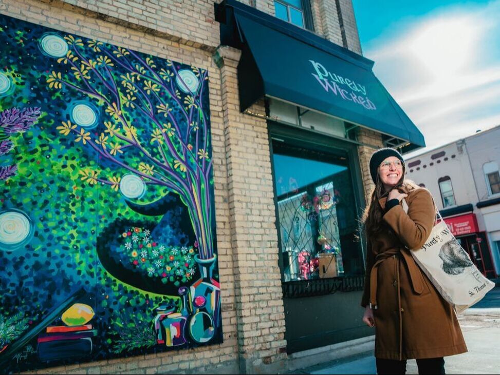 a woman holding a shopping tote in front of a downtown mural