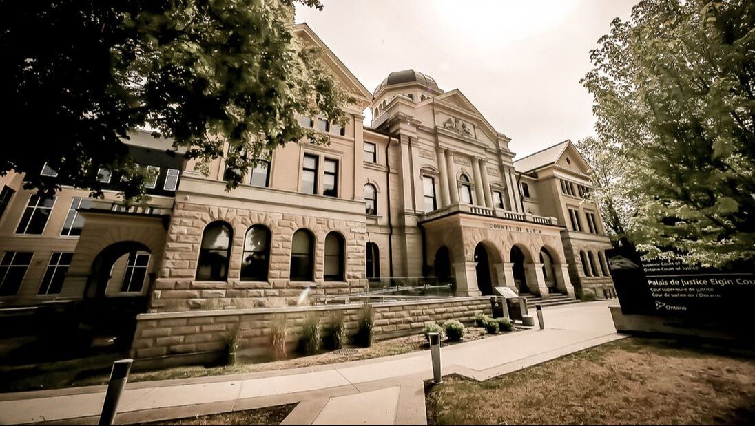 Here is an angled shot of the Court House. To the left and right of the photo are green leaves of trees nearby. Just off centre to the right of the photo is the three arch entry way to the courthouse. By the roof of the courthouse are a unicorn and another animal facing off. Above this is a circular dome like area of the courthouse with a black roof. The bottom portion of the courthouse is covered in large beige stone. There are many windows both rectangular and rounded throughout the building. 