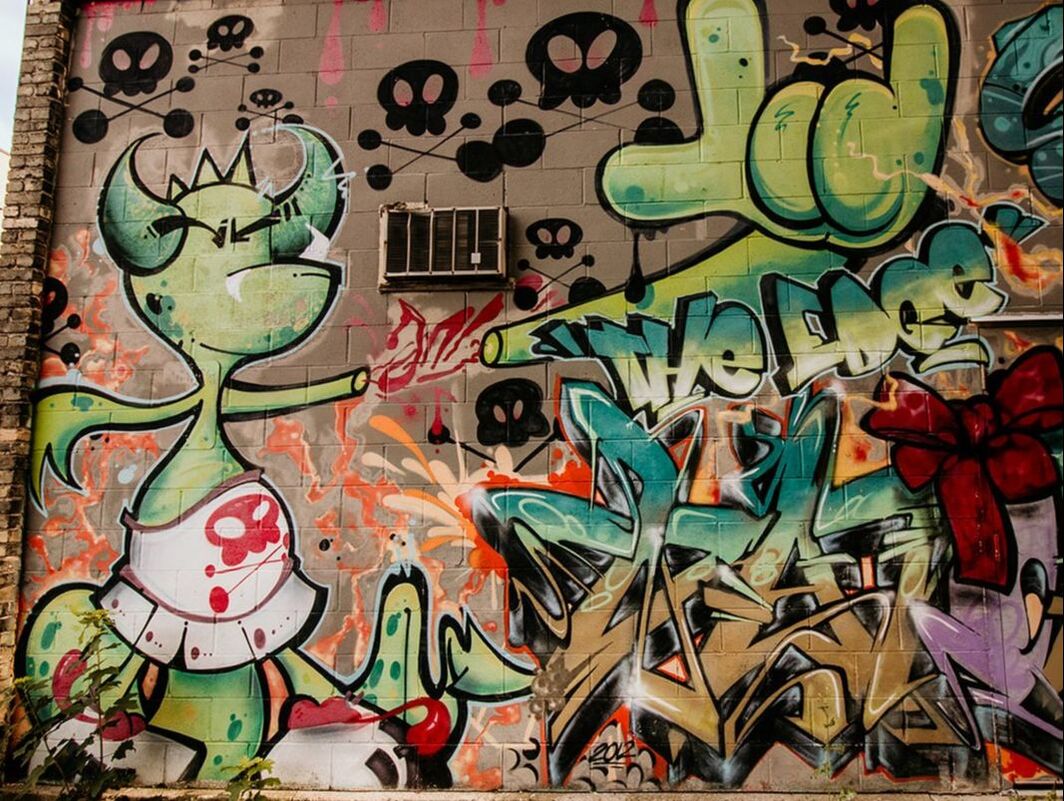 This is an image of a graffiti like mural. To the bottom left of the mural is a cartoon like dragon with a white skirt with a red skull on it. To the right bottom corner there are graffiti symbols in the brown, turquoise, burgundy, and light green. At the top are black skulls with red drips from the roof.