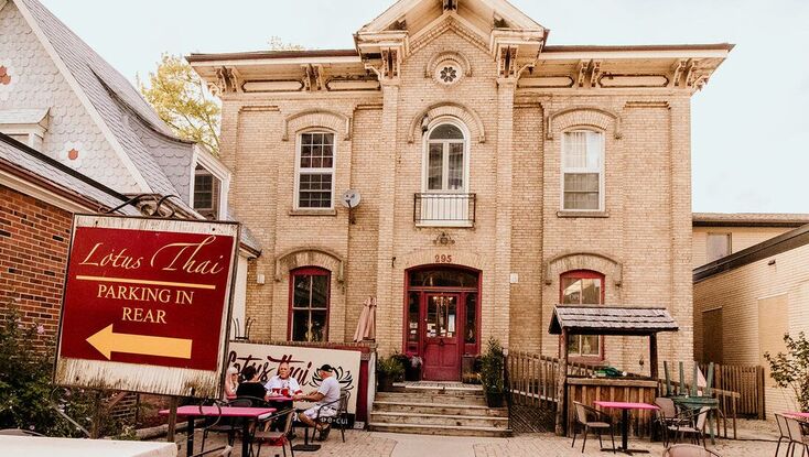 Here is Lotus Thai restaurant. Straight ahead is the historic beige bricked building. The front door is red with windows on either side. The windows are also framed in red. The second floor of the house has three windows, with the one in the middle having a black balcony bar in front. Below the house is a small front deck with a ramp to the right. In front of the deck is a table of four sitting on the patio. At the very front of the picture is a sign that says 'Lotus Thai Parking in Rear' with an arrow pointing left.