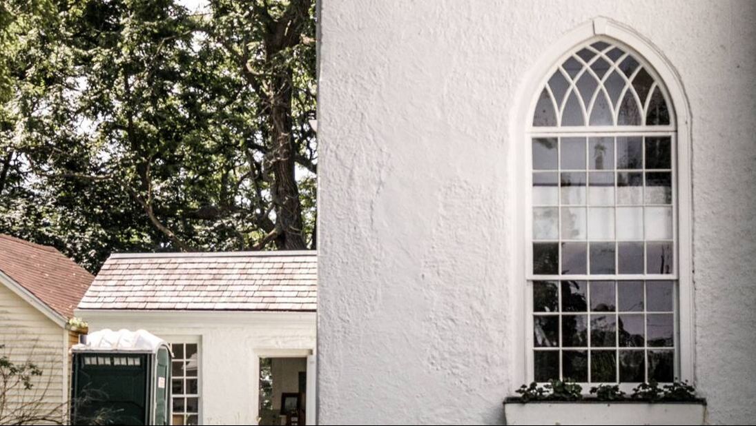 Here is Old St. Thomas Church. This photo shows off one of the corners of the church. The building is completely white. The windows are white paned arched windows. Below the window is a flower box with green plants. To the left of the image is a door which is open leading inside the church.