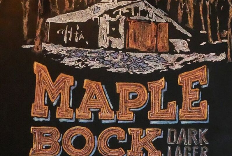 This is a drawing of the maple shack in Springwater. The top portion is the maple shack in the winter. The roof is white, and to the right of the shack is a brown fence. Behind the shack are brown trees representing the forest behind it. In front of the shack is snow mixed with a few blue patches to represent ice. Below the snow are big bold brown letters saying 'Maple Bock' and beside 'Bock' are smaller letters saying 'Dark Lager'. 