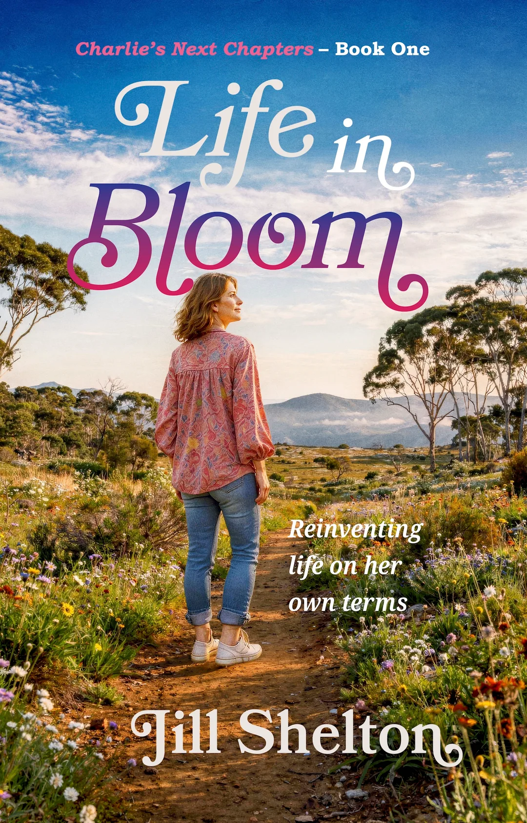 Cover of Life in Bloom by Jill Shelton, Book One in Charlie’s Next Chapters. A woman with shoulder-length light brown hair stands on a dirt path bordered by colourful wildflowers, her body facing away as she looks to the right. She wears a pink patterned blouse, rolled-up blue jeans and white trainers. Eucalyptus trees frame an open countryside landscape with distant mountains beneath a blue sky streaked with white clouds. The title appears in large flowing white, purple and pink lettering. The tagline reads, “Reinventing life on her own terms.”