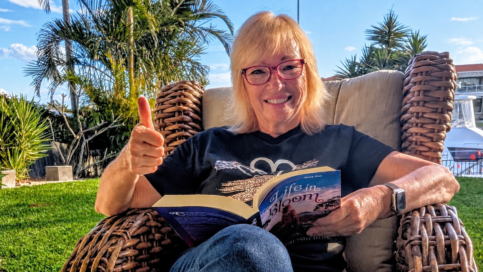 Author Jill Shelton sitting outdoors in a wicker chair, smiling and giving a thumbs-up while holding an open copy of her book, Life in Bloom.