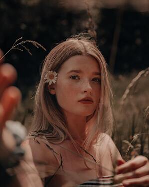 Young woman with blonde hair and a daisy behind her ear looking into the camera. This illustrates that people-pleasing and codependency is common in young women.