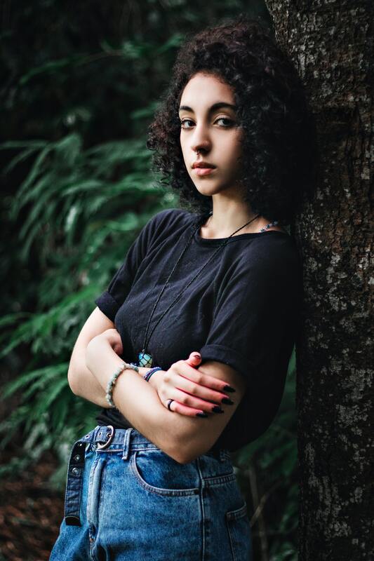 Young woman with curly dark hair leaning against a tree, arms crossed. This illustrates that Sacred Circle Holistic Healing offers online therapy in Kansas for anxiety, low self-esteem, and codependency.