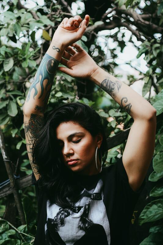 Woman with brown skin, black hair, and tattoos on her arms dancing with her eyes closed. This represents that Sacred Circle Holistic Healing offers online therapy in Kansas for young woman struggling with anxiety and low self-esteem.